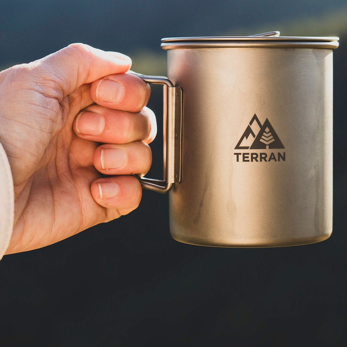Hand holding a metallic mug with 'Terran' logo against a blurred natural background