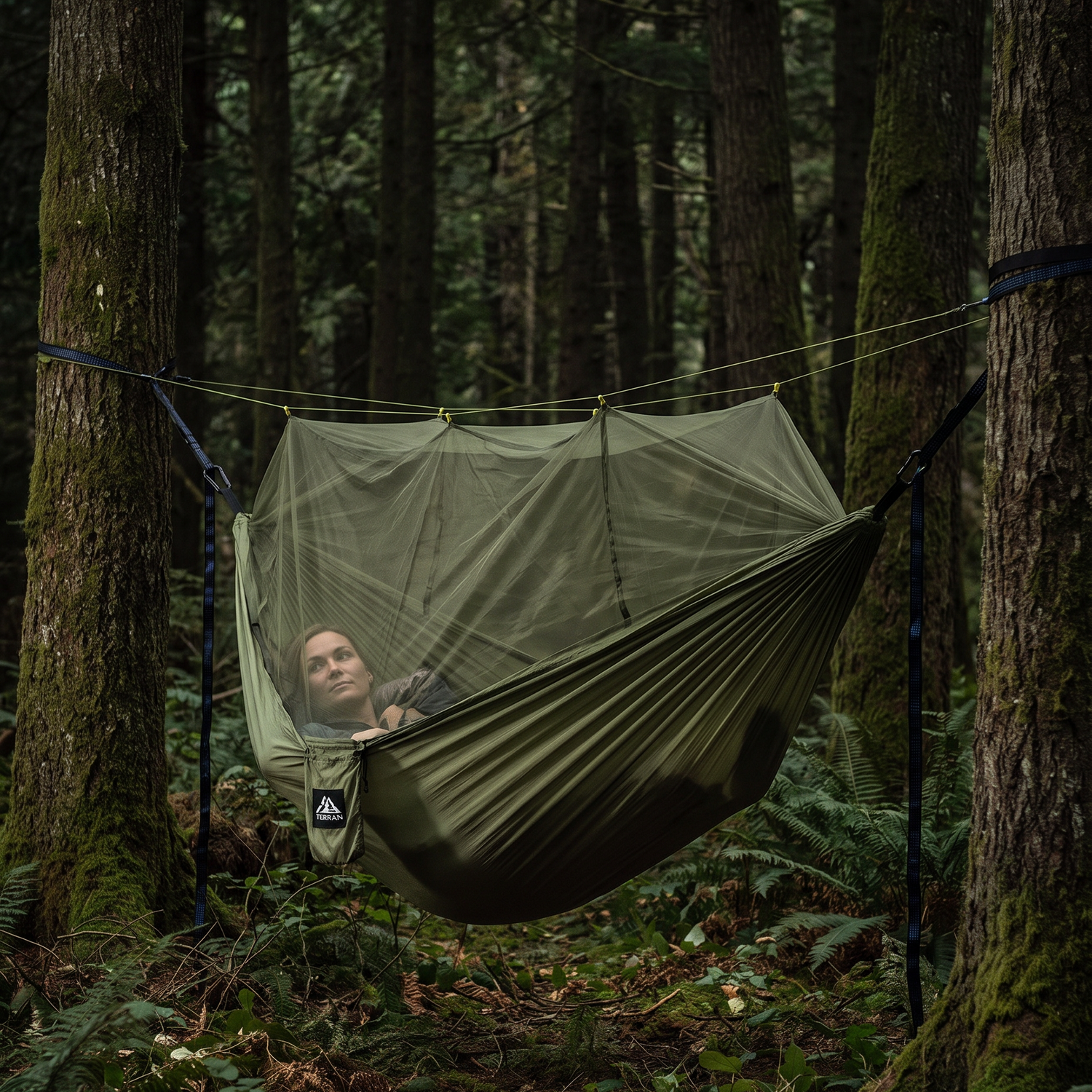 An olive green camping hammock with an attached mosquito net enclosure, suspended between trees in a dense forest. A person is lying inside the hammock, looking up.