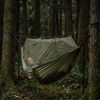An olive green camping hammock with an attached mosquito net enclosure, suspended between trees in a dense forest. A person is lying inside the hammock, looking up.